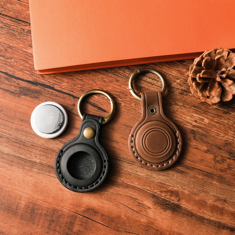 May include: A white AirTag and two leather keychains in black and brown. The keychains have gold-colored hardware and are designed to hold the AirTag. The items are on a wooden surface.