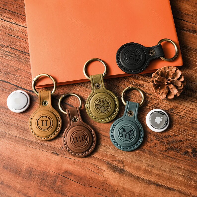 May include: A collection of leather keychains in various colors, including brown, olive green, and black, each with a gold-colored keyring. Some keychains feature embossed initials or designs. A white Apple AirTag is also present.