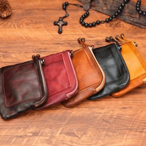 May include: Six leather zippered pouches in various colors, including brown, red, and black, arranged in a row on a wooden surface.