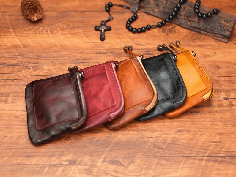 May include: Six leather zippered pouches in various colors, including brown, red, and black, arranged in a row on a wooden surface.