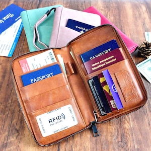 May include: A brown leather travel wallet with a zipper closure, open to reveal multiple pockets holding passports, credit cards, and boarding passes. The wallet is designed to protect against RFID theft. Other travel wallets are visible in the background.