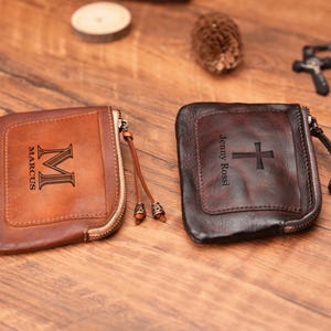 May include: Two personalized leather zipper pouches. One is brown with the name "Marcus" and the initial "M" engraved. The other is black with the name "Jenny Rossi" and a cross engraved.