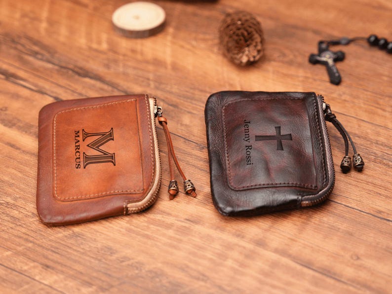 May include: Two personalized leather zipper pouches. One is brown with the name "Marcus" and the initial "M" engraved. The other is black with the name "Jenny Rossi" and a cross engraved.