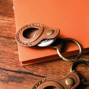 May include: A brown leather keychain holder with a metal ring and a gold-colored button. The holder is stitched around the edges and holds a white circular object. The background includes an orange notebook and a wooden surface.