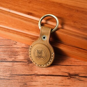May include: A tan leather keychain holder with a metal ring. The holder features an embossed owl design and white stitching around the edges. The keychain is resting on a wooden surface.