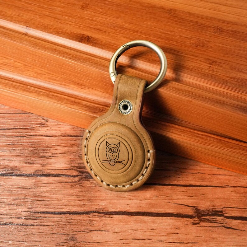 May include: A tan leather keychain holder with a metal ring. The holder features an embossed owl design and white stitching around the edges. The keychain is resting on a wooden surface.