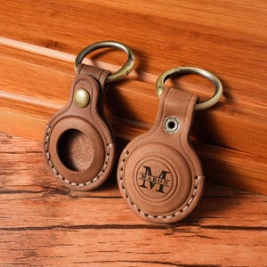 May include: Two brown leather keychains with metal rings. One keychain has the word "MABON" and the letter "M" engraved on it. The other keychain has a circular opening. Both keychains have a stitched border and a metal button.