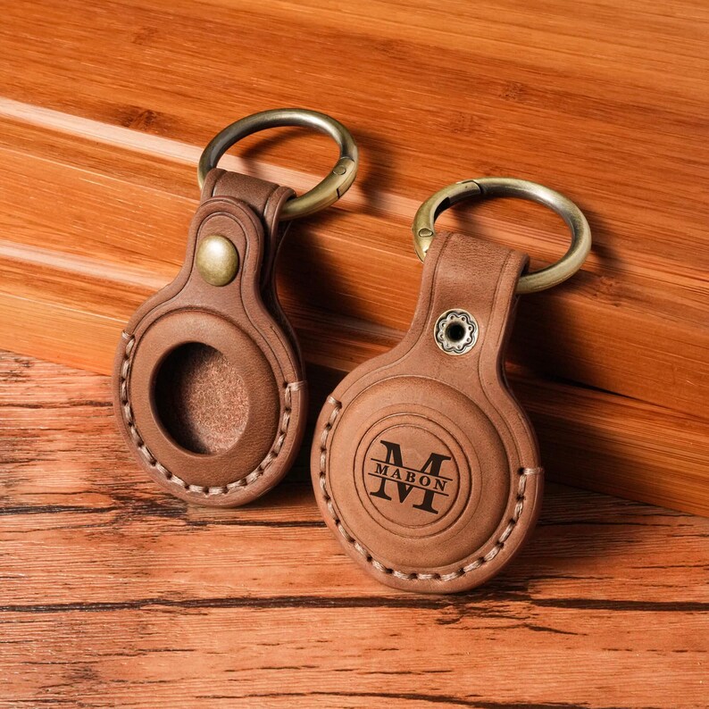 May include: Two brown leather keychains with metal rings. One keychain has the word "MABON" and the letter "M" engraved on it. The other keychain has a circular opening. Both keychains have a stitched border and a metal button.