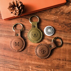 May include: Three personalized leather keychains with gold-colored hardware, each featuring a different initial or monogram. One is olive green, and two are brown. A white circular tracker is also present. The items are arranged on a wooden surface.