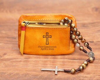May include: A brown leather wallet with a zipper closure and a cross engraved on the front. The wallet is personalized with the text "First Communion April 1, 2025". A brown beaded rosary with a silver cross charm is attached to the wallet.