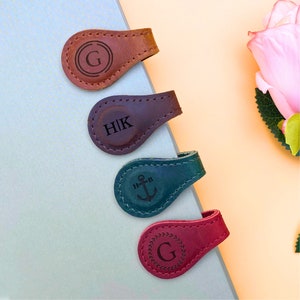 Könnte beinhalten: Vier Leder-Lesezeichen mit verschiedenen gravierten Designs. Die Lesezeichen sind braun, dunkelviolett, dunkelblau und burgunderrot. Die Designs beinhalten einen Kreis mit dem Buchstaben G, die Buchstaben HIK, einen Anker und einen Kreis mit dem Buchstaben G, umgeben von einem Kranz.