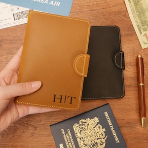 May include: Two passport holders, one brown and one black, with a monogrammed "HIT" on the brown holder. The holders are made of leather and have a flap closure. A British passport is visible in the background.