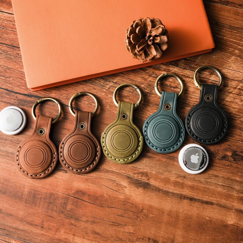 May include: A collection of leather keychains in various colors, including brown, olive green, teal, and black, with gold-colored key rings. The keychains are arranged on a wooden surface, alongside an orange notebook and a pine cone.