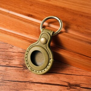 May include: Olive green leather key fob with a brass-colored metal ring. The fob has a circular opening and a button closure. The leather is stitched with a lighter green thread. The key fob is designed to hold a small electronic device.