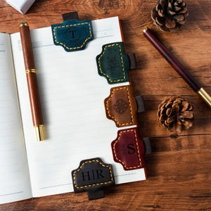 Op de afbeelding: Een notitieboek met verschillende leren bladwijzers in diverse kleuren, waaronder teal, groen, bruin en bordeauxrood. Elke bladwijzer heeft een uniek ontwerp of initiaal. Een houten pen en dennenappels zijn ook zichtbaar.