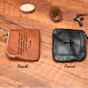 May include: A brown leather pouch with a zipper closure and a quote engraved on the back. The front of the pouch is black leather with a cross design.
