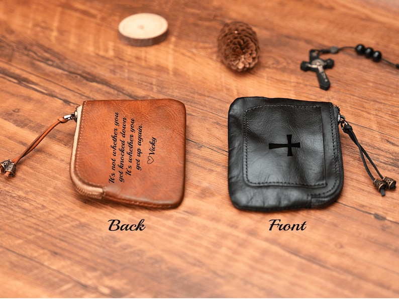 May include: A brown leather pouch with a zipper closure and a quote engraved on the back. The front of the pouch is black leather with a cross design.