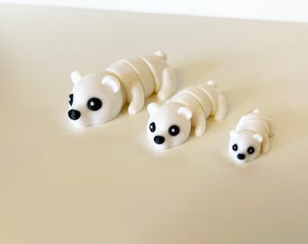 May include: Three white polar bear figurines with black eyes and noses are arranged on a light beige surface. The bears are in a lying down position, with the largest bear at the top and the smallest at the bottom. The bears are a cute, whimsical design.