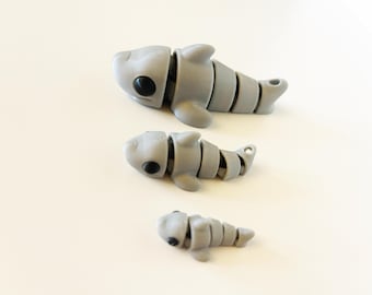 May include: Three gray articulated shark toys with black eyes are arranged on a white surface. The sharks are segmented, allowing for movement. The largest shark is at the top, followed by a medium and a small shark.