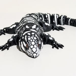 May include: A black and white lizard figurine with a detailed pattern. The reptile is lying flat, showcasing its textured skin and realistic features. The figurine has small, dark eyes and is set against a plain white background.