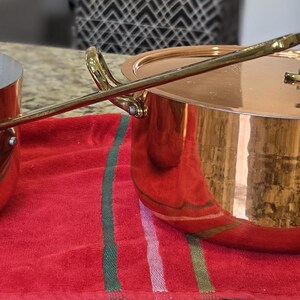 May include: Two copper cooking pots with brass handles and lids sit on a red cloth. The larger pot is open, revealing a silver interior. The smaller pot has a lid and a long brass spoon. The pots are reflective and gleam.