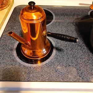 May include: A polished copper-colored coffee pot with a cylindrical body, a short angled spout, and a black handle. The pot features a matching lid with a small black knob on top, reflecting light on its smooth, metallic surface.