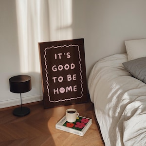 May include: A framed brown wall art with the text "IT'S GOOD TO BE HOME" in pink, a black table lamp, a coffee cup, and a book with colorful flowers. The items are arranged on a wooden floor next to a bed with white bedding.
