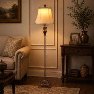 May include: A floor lamp with a tall, slender, dark bronze-toned metal stand featuring an ornate, bulbous decorative element below a flared, off-white fabric lampshade that emits a warm, soft glow, resting on a square, detailed base.