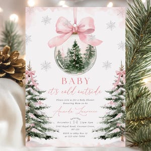 May include: A baby shower invitation with a winter theme. The design features a pink bow, a glass ornament with a winter scene, and two Christmas trees. The text reads "BABY it's cold outside".