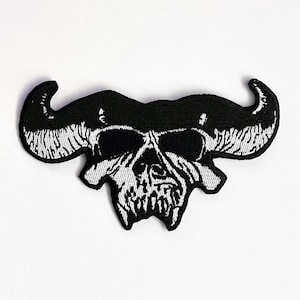 Op de afbeelding: Zwart-witte geborduurde patch van een schedel met hoorns. De schedel heeft een dreigende uitdrukking met scherpe tanden en een gefronste wenkbrauw.