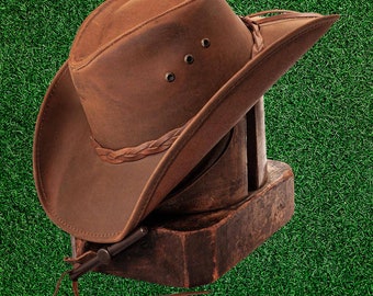 Puede incluir: Un sombrero vaquero de cuero marrón con una banda trenzada y una correa de barbilla de cuero. El sombrero está sentado en una forma de sombrero de madera.