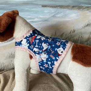 May include: A blue dog harness with a patriotic print featuring white dogs, bones, stars, and fireworks. The harness has a red and white trim and is designed to fit a dog. The dog in the image is white and brown.