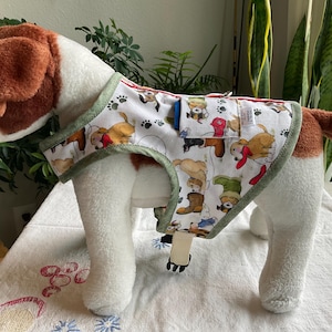 May include: A plush dog toy wearing a patterned vest. The vest features a white background with colorful illustrations of dogs in various poses, paw prints, and a green trim. The dog toy has brown and white fur.