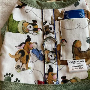 May include: Dog harness with a playful puppy print on a white background. The harness features a green trim and a small pocket. The design includes cartoon dogs, paw prints, and a blue shoe. The text "WILLIE BARKS" is visible.