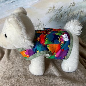 May include: A white plush dog wearing a colorful frog-patterned vest. The vest features a vibrant design with frogs in various colors, including blue, orange, and purple, with a green trim. The dog is on a soft, tan surface.