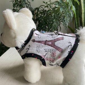 May include: White dog harness with a black trim and a Paris-themed print. The print features the Eiffel Tower in pink, along with other Parisian buildings and clouds. The harness has a metal buckle and a D-ring for leash attachment.