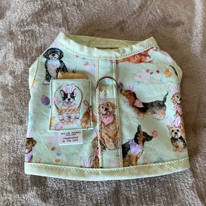 May include: A light green dog harness featuring an Easter-themed print with various dog breeds wearing bunny ears and surrounded by Easter eggs. The harness includes a pocket and a metal ring for leash attachment. The text "WILLIE BARKS Handcrafted In The USA" is visible.