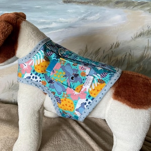 May include: A dog harness with a colorful tropical print featuring pineapples, flowers, and a cartoon character. The harness has a teal background with blue trim and a small pocket. The dog is a stuffed animal with brown and white fur.