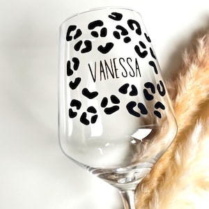 Op de afbeelding: Een helder wijnglas met een zwart luipaardprintontwerp en de naam "VANESSA" in een dun, zwart lettertype. Het glas staat op een witte achtergrond met gedroogd, bruinachtig decoratief gras.