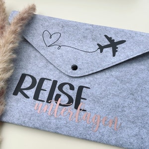 Könnte beinhalten: Graue Filz-Reiseunterlagen-Tasche mit schwarzem Flugzeug- und Herz-Design. Das Wort "REISE" ist schwarz gedruckt, "unterlagen" in Roségold-Schrift. Mit einem Druckknopfverschluss.
