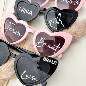 Op de afbeelding: Vijf paar hartvormige zonnebrillen met verschillende tekst op elke lens. De zonnebrillen zijn zwart en roze. De tekst op de lenzen is "Ninas JGA", "21-06-24", "NINA", "Team", "Braut", "BRAUT" en "Luisa".