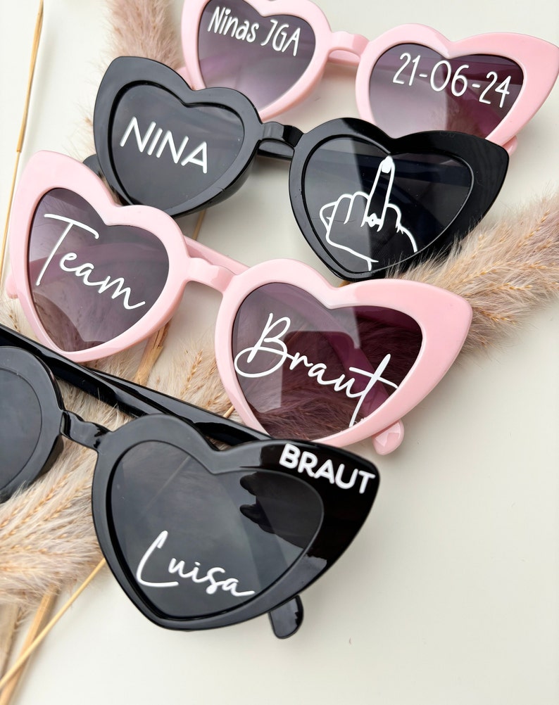 Op de afbeelding: Vijf paar hartvormige zonnebrillen met verschillende tekst op elke lens. De zonnebrillen zijn zwart en roze. De tekst op de lenzen is "Ninas JGA", "21-06-24", "NINA", "Team", "Braut", "BRAUT" en "Luisa".