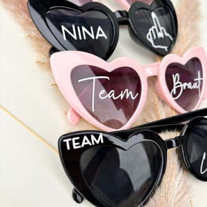 Op de afbeelding: Vier paar hartvormige zonnebrillen met verschillende tekst op elke lens. De zonnebrillen zijn zwart en roze. De tekst op de lenzen luidt "Ninas JGA 21-06-24", "NINA", "Team" en "Braut".