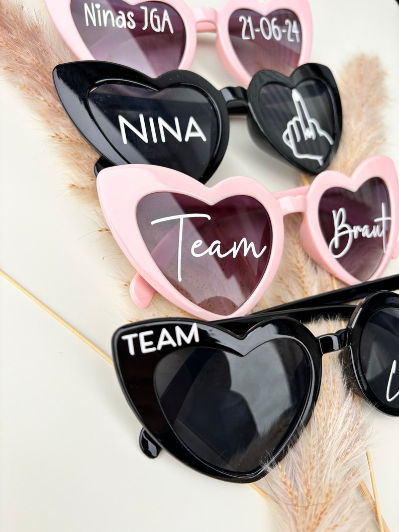 Op de afbeelding: Vier paar hartvormige zonnebrillen met verschillende tekst op elke lens. De zonnebrillen zijn zwart en roze. De tekst op de lenzen luidt "Ninas JGA 21-06-24", "NINA", "Team" en "Braut".