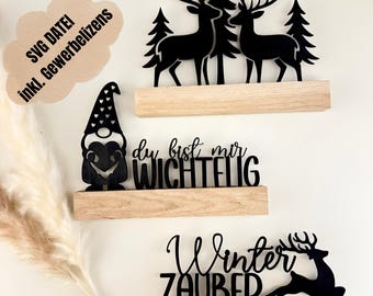 Könnte beinhalten: Drei Holzregale mit schwarzen Laser-Cut-Dekorationen. Das obere Regal zeigt Hirsche und Kiefern. Das mittlere Regal trägt den Text "du bist mir WICHTIG". Das untere Regal zeigt "Winter ZAUBER" mit einer Hirschsilhouette. Eine Sprechblase mit Text ist oben links.