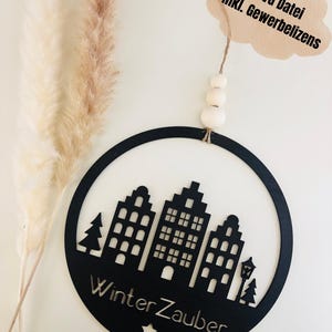 Könnte beinhalten: Schwarze kreisförmige Wanddekoration mit einem ausgeschnittenen Design von Gebäuden, Bäumen und einer Straßenlaterne. Die Worte "Winter Zauber" sind unten ausgeschnitten. Ein Anhänger mit Text hängt oben. Pampasgras im Hintergrund.