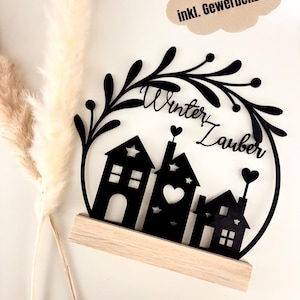 May include: Black wooden winter-themed decoration featuring three stylized houses with heart and star cutouts, set against a circular wreath with the words "Winter Zauber". The houses sit on a light wood base. Text in a cloud reads "SVG Datei inkl. Gewerbelizenzens".