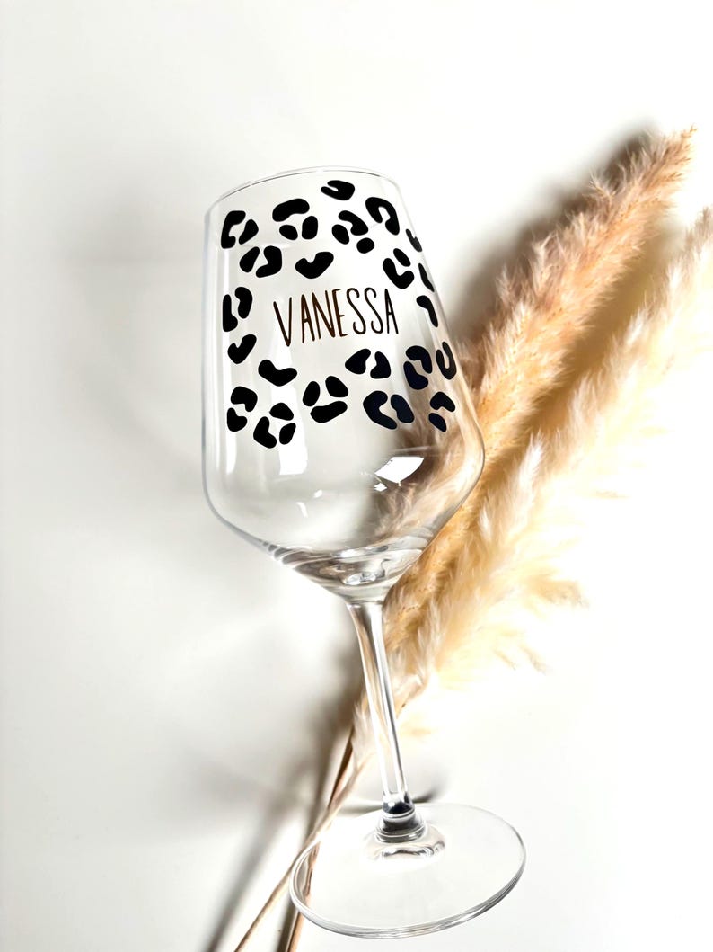Op de afbeelding: Een helder wijnglas met een zwart luipaardprintontwerp en de naam "VANESSA" in een dun, zwart lettertype. Het glas staat op een witte achtergrond met gedroogd, bruinachtig decoratief gras.