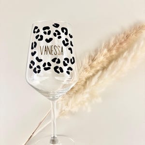 Op de afbeelding: Een helder wijnglas met een luipaardprint en de naam "Vanessa" in gouden letters. Het glas heeft een lange steel en een brede kom. Gedroogde decoratieve planten zijn op de achtergrond te zien.