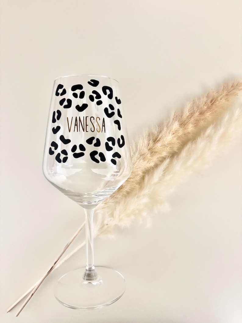 Op de afbeelding: Een helder wijnglas met een luipaardprint en de naam "Vanessa" in gouden letters. Het glas heeft een lange steel en een brede kom. Gedroogde decoratieve planten zijn op de achtergrond te zien.